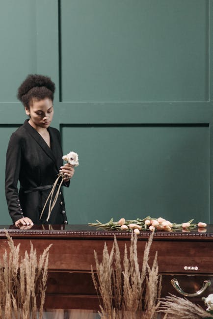 A young woman in a black dress stands beside a wooden casket decorated with a floral arrangement featuring soft pink roses and peach-colored blooms, with green foliage. She holds a small bouquet of wh