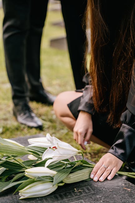 A person dressed in black kneels on the ground, arranging a floral tribute of white lilies and long green leaves on a memorial stone, with a grief-stricken hand gently touching the flowers. In the bac