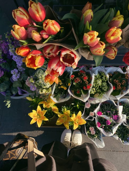 A top-down view of a floral display featuring a variety of fresh flowers arranged in a market setting; prominently, a bouquet of pink and yellow tulips with green stems and leaves is placed on top, su