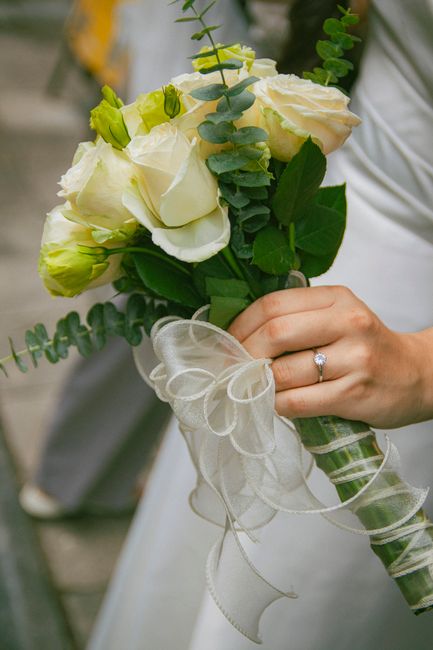 A close-up of a bride's hand holding a delicate bouquet arranged with white roses, light green eustoma buds, and sprigs of eucalyptus, wrapped in sheer white ribbon. The bouquet's fresh flowers displa