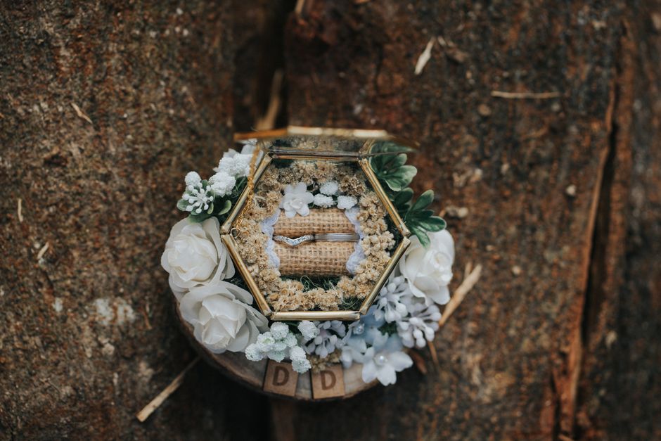 A delicate hexagonal glass ring box with gold framing, containing a wedding band, is encircled by a floral arrangement of soft white roses, small white blossoms, and green foliage. The box and flowers
